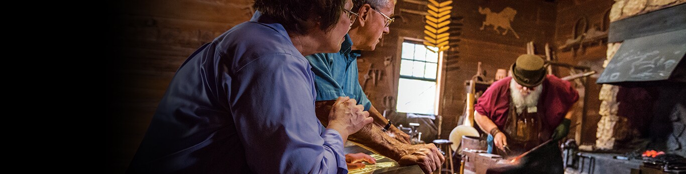 Äldre par som tittar på medan smeden arbetar på en del till en metallhammare på den historiska platsen i Grand Detour, Illinois, USA