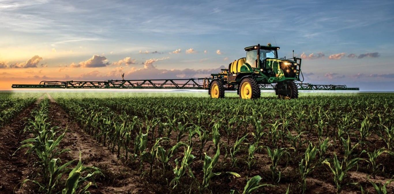 En John Deere besprutare som arbetar på en sädesåker med solnedgången i bakgrunden