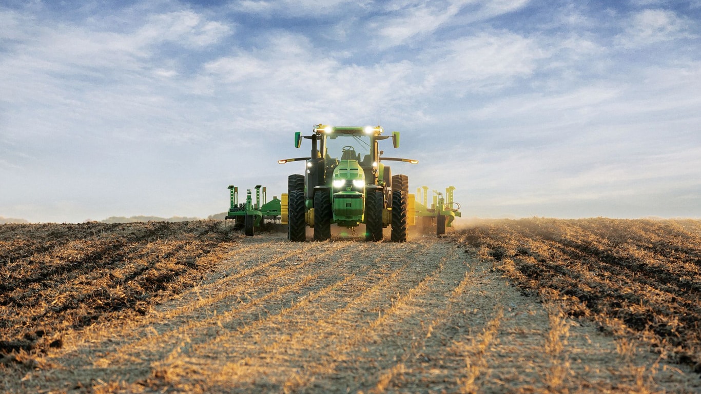 Självkörande traktor från John Deere drar jordbearbetningsutrustning genom ett öppet fält.