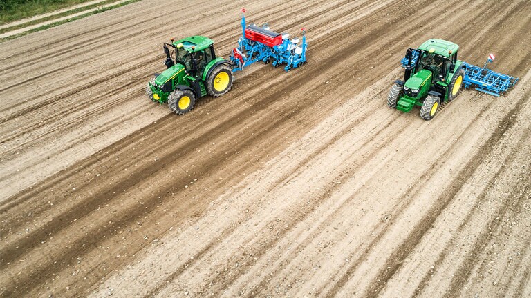 Tv&aring; John Deere 6M-traktorer p&aring; ett kalt jordf&auml;lt. Den ena drar en Monsomen-planteringsmaskin. Den andra drar en Lemken-fr&ouml;b&auml;ddssk&ouml;rdetr&ouml;ska