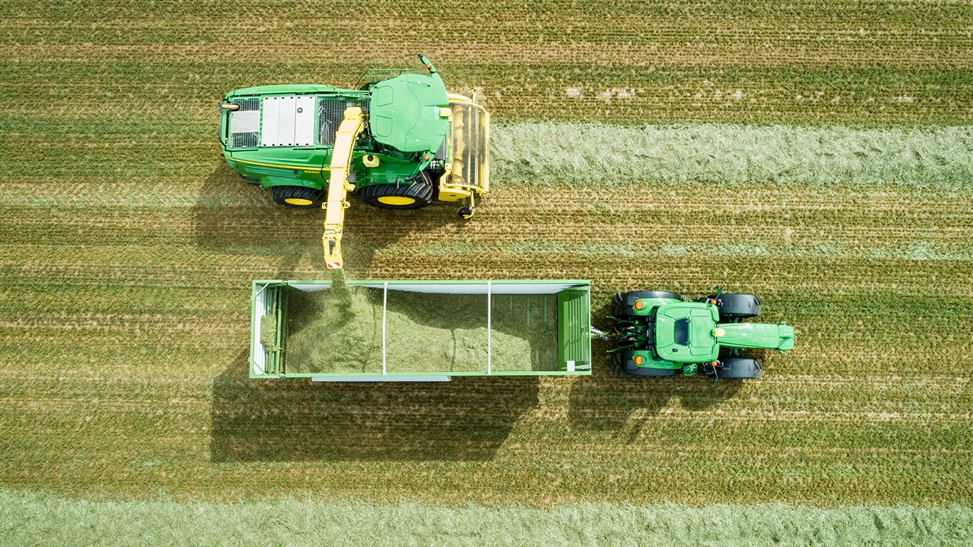 Flygfoto av John Deere 8000-seriens sj&auml;lvg&aring;ende exakthack med en gr&auml;spickup som lastar silage i en vagn som dras av en John Deere-traktor