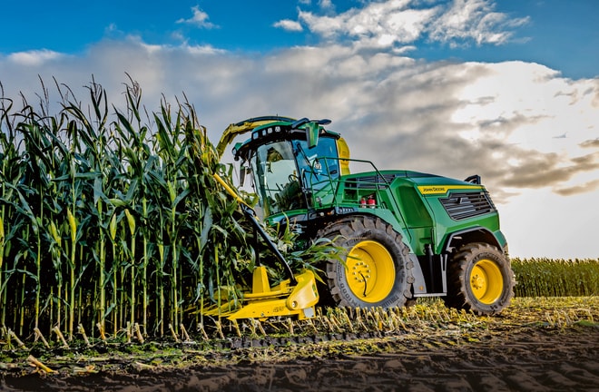 John Deere exakthack skördar majs på ett fält under blå himmel med moln.